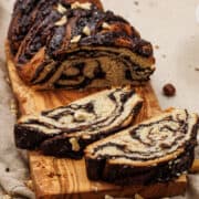 A sliced loaf of chocolate hazelnut babka rests on a wooden cutting board. The bread boasts a swirled pattern of indulgent chocolate and dough, crowned with chopped nuts. In the background, a beige tablecloth is adorned with scattered crumbs and a few hazelnuts.