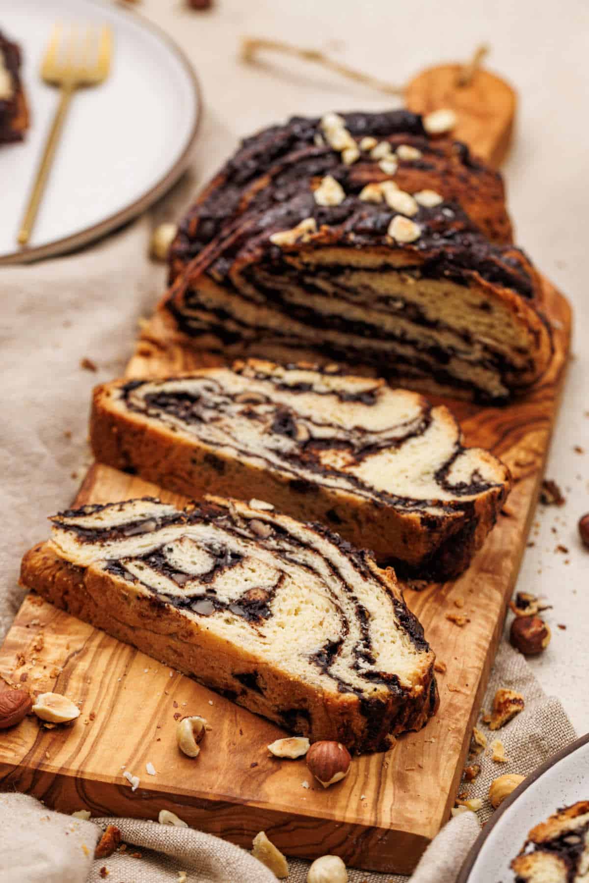 A wooden board holds a sliced chocolate hazelnut babka, showcasing its marbled pattern. Hazelnuts are scattered around, while part of a plate with a fork peeks in the background. The babka boasts a golden crust and rich chocolate layers.