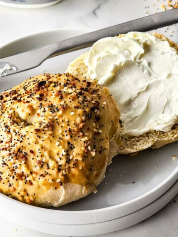 A split everything bagel sits on a white plate, one half generously spread with cream cheese. A butter knife rests beside it, seeds scattered around hinting at the flavorful dance of everything bagels.