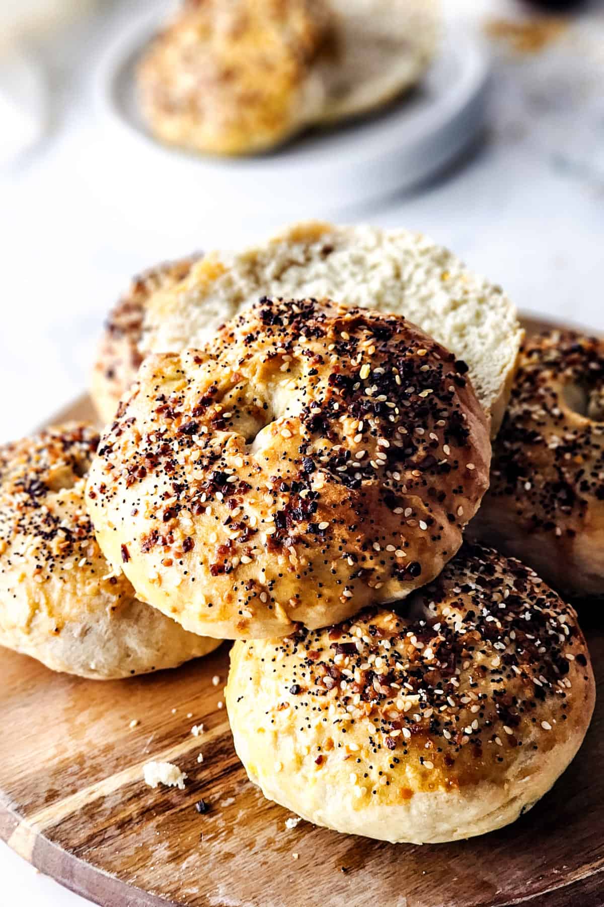 A wooden board topped with everything bagels, their crusts speckled with mixed seeds and seasonings. One bagel is sliced in half, revealing the soft interior. A blurred plate with another sliced everything bagel adds depth to the background.