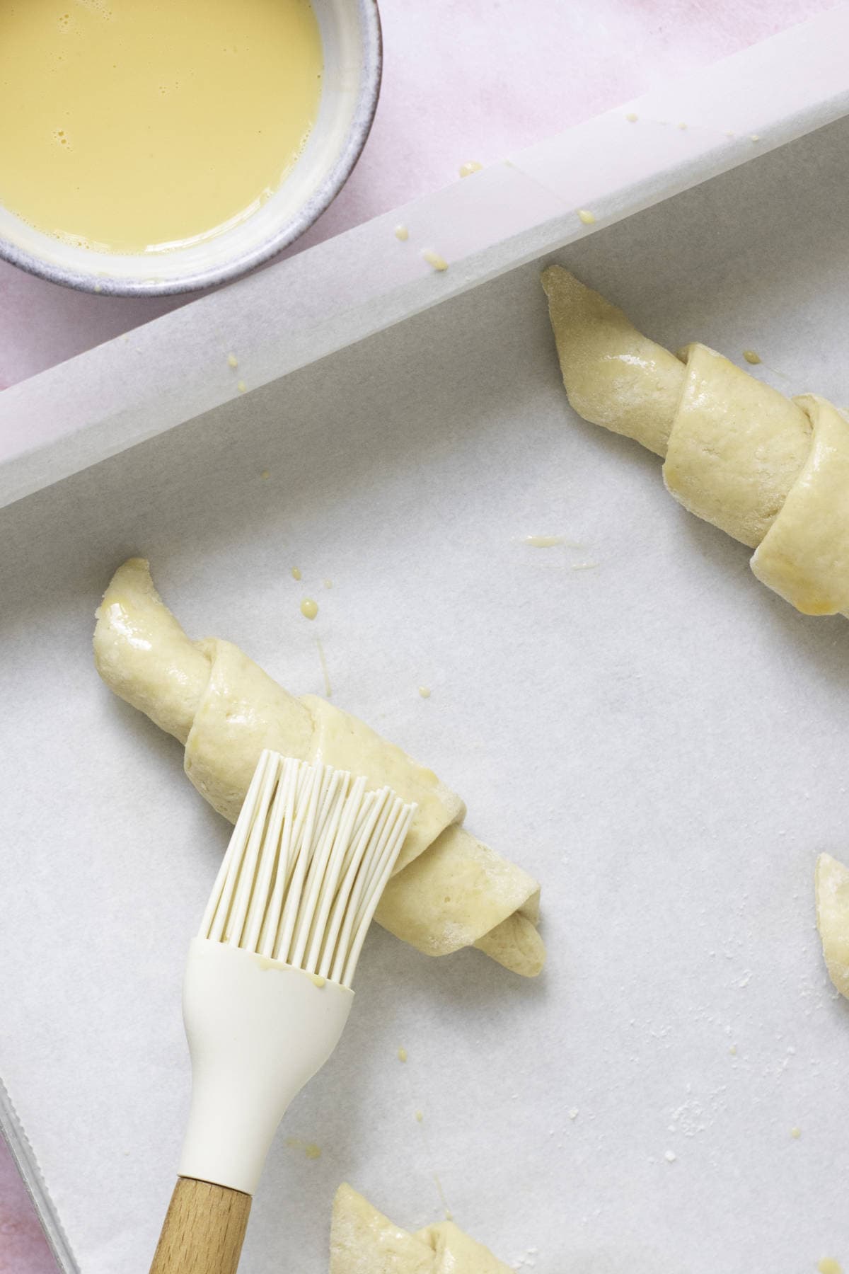 A baking tray lined with parchment paper holds unbaked, homemade croissants. A white pastry brush with some egg wash rests on one croissant, promising a golden finish. A small bowl with more egg wash peeks from the corner, perfecting this fool-proof croissant preparation.