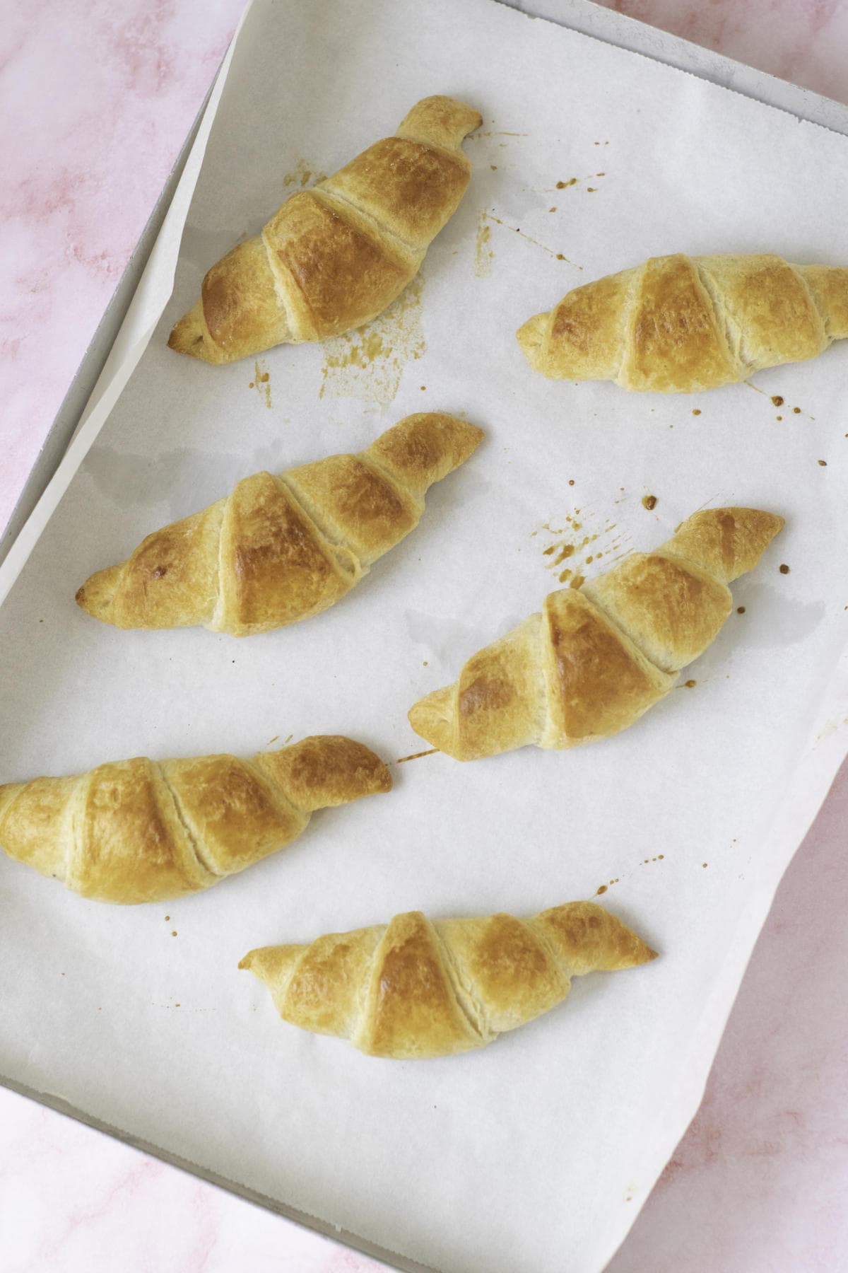Six homemade croissants, golden-brown and flaky, sit on a parchment-lined baking tray, resting on a pink marbled surface.