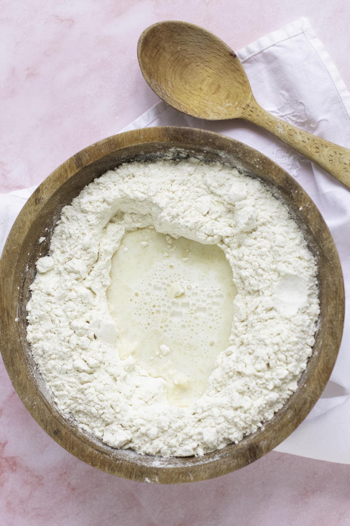A wooden bowl filled with flour, ready for homemade croissants, features a well in the center containing liquid. The bowl rests on a white cloth, with a wooden spoon nearby on a pink surface.