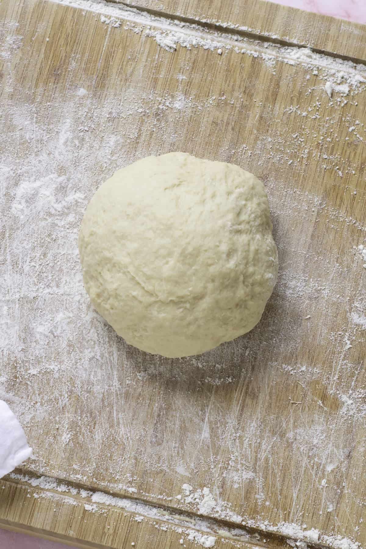 A ball of dough destined for fool-proof croissants sits on a floured wooden cutting board. The surface around the dough is lightly dusted with flour, and a white cloth is partially visible in the corner.