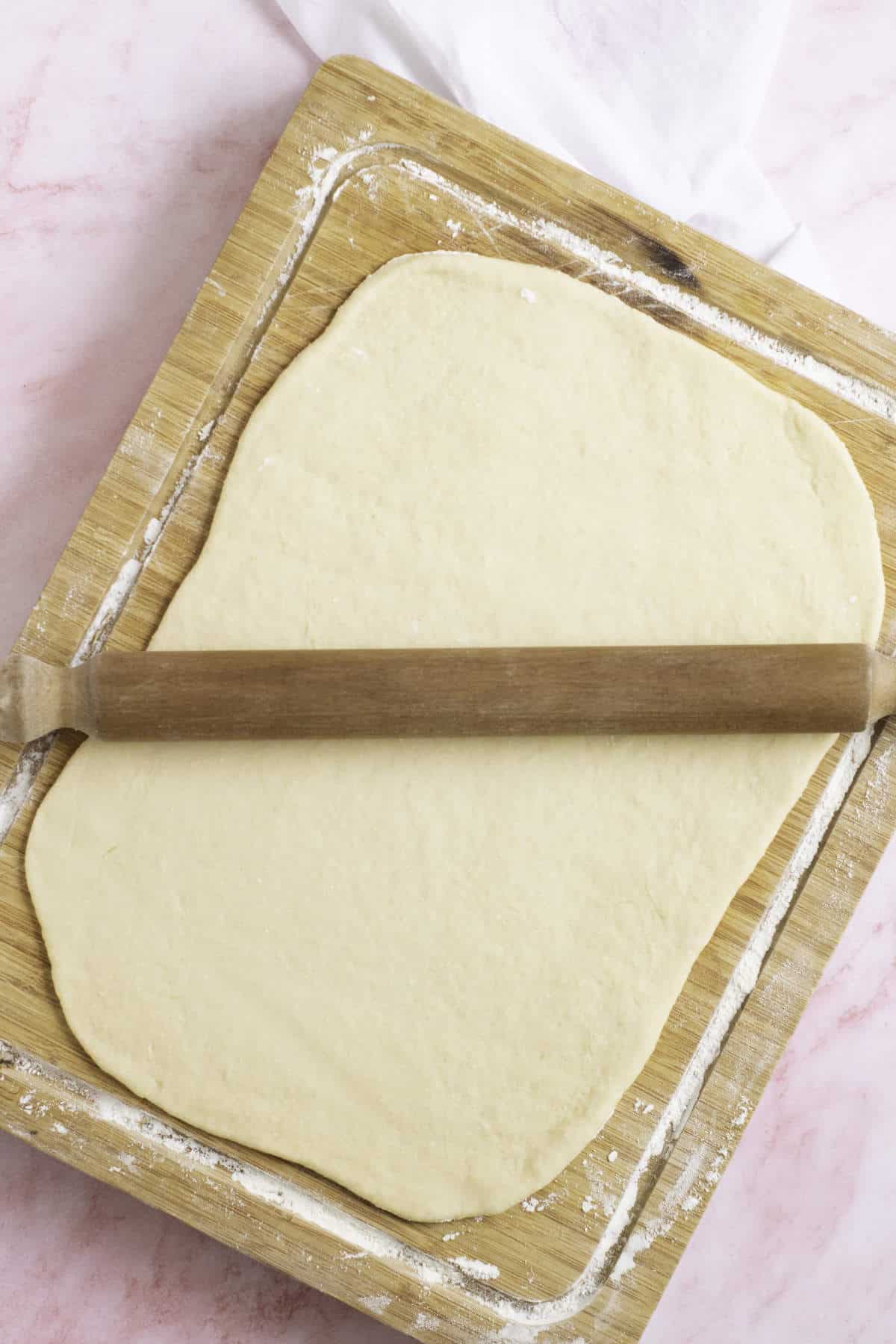 A wooden rolling pin rests on raw, rolled-out dough atop a floured wooden cutting board, perfect for crafting homemade croissants. The board sits on a light pink marbled surface, with a white cloth partially visible in the corner.