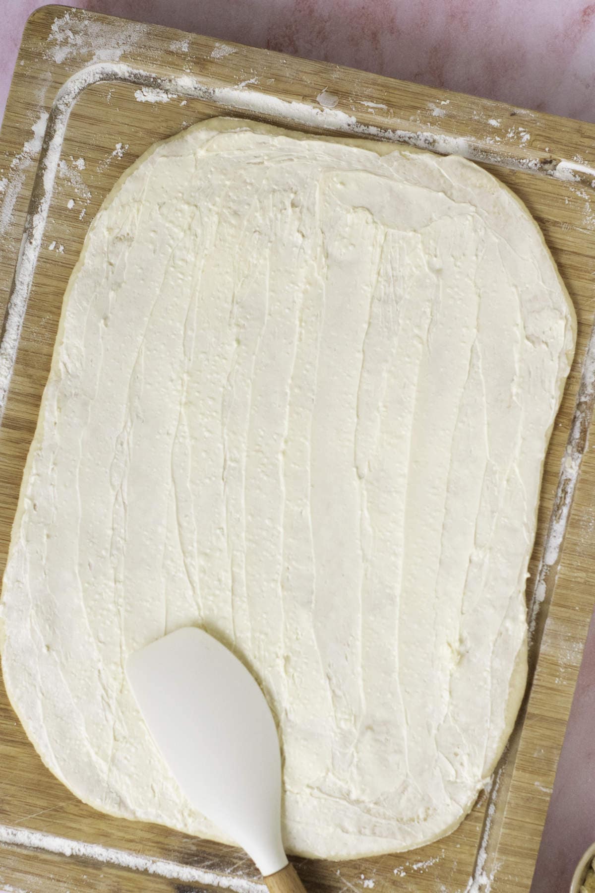 A rectangular piece of dough is rolled out on a floured wooden surface. A white spatula spreads cream cheese over it, laying the foundation for fool-proof croissants. The background is slightly blurred with a pinkish hue.