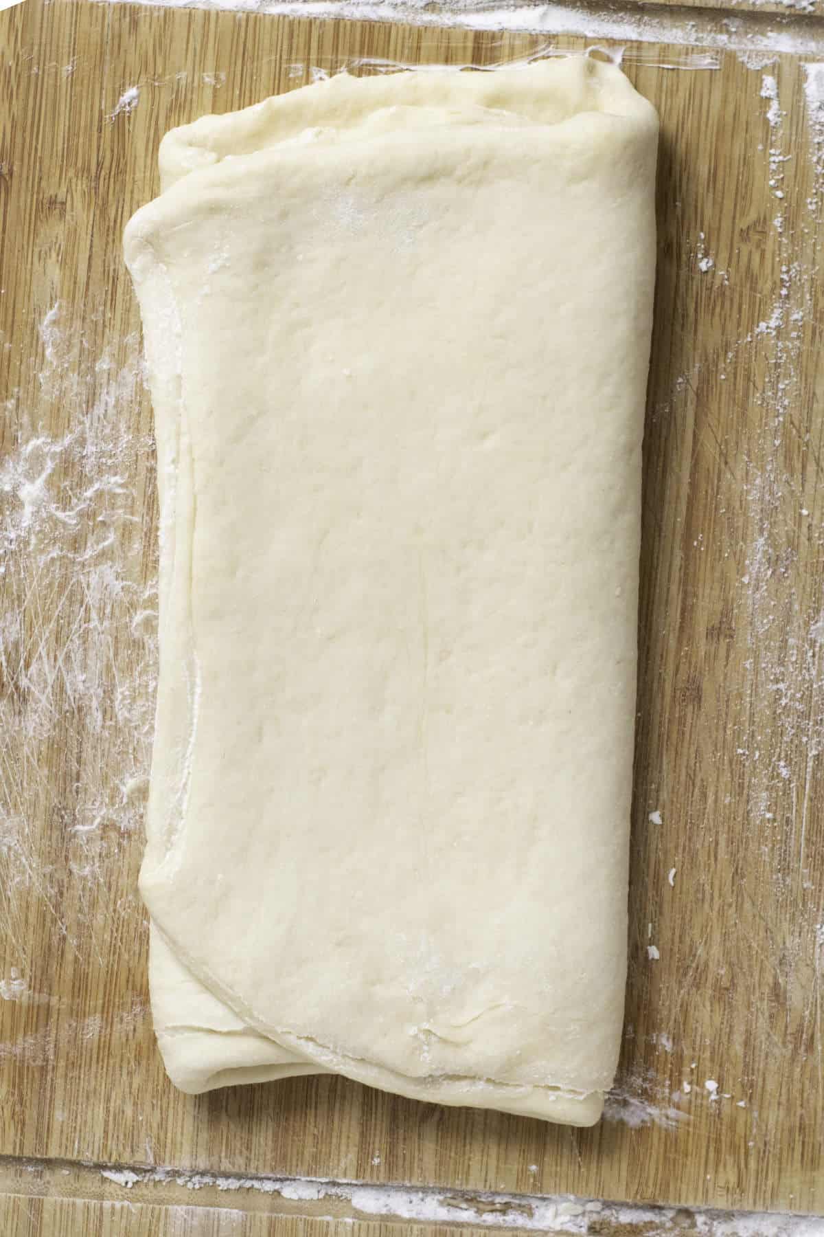 A folded rectangle of raw puff pastry dough sits on a wooden surface, lightly dusted with flour-an inviting start for fool-proof homemade croissants.