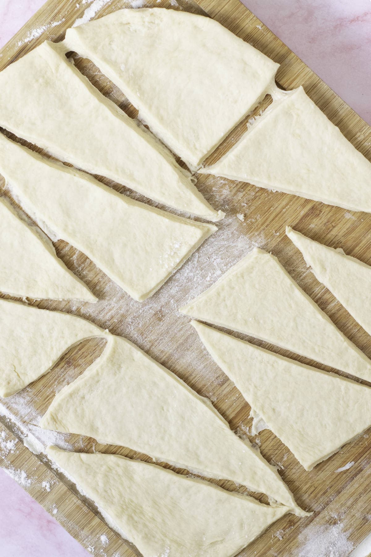 Triangle-shaped pieces of rolled dough on a floured wooden surface, these homemade croissants are fool-proof and ready for baking.