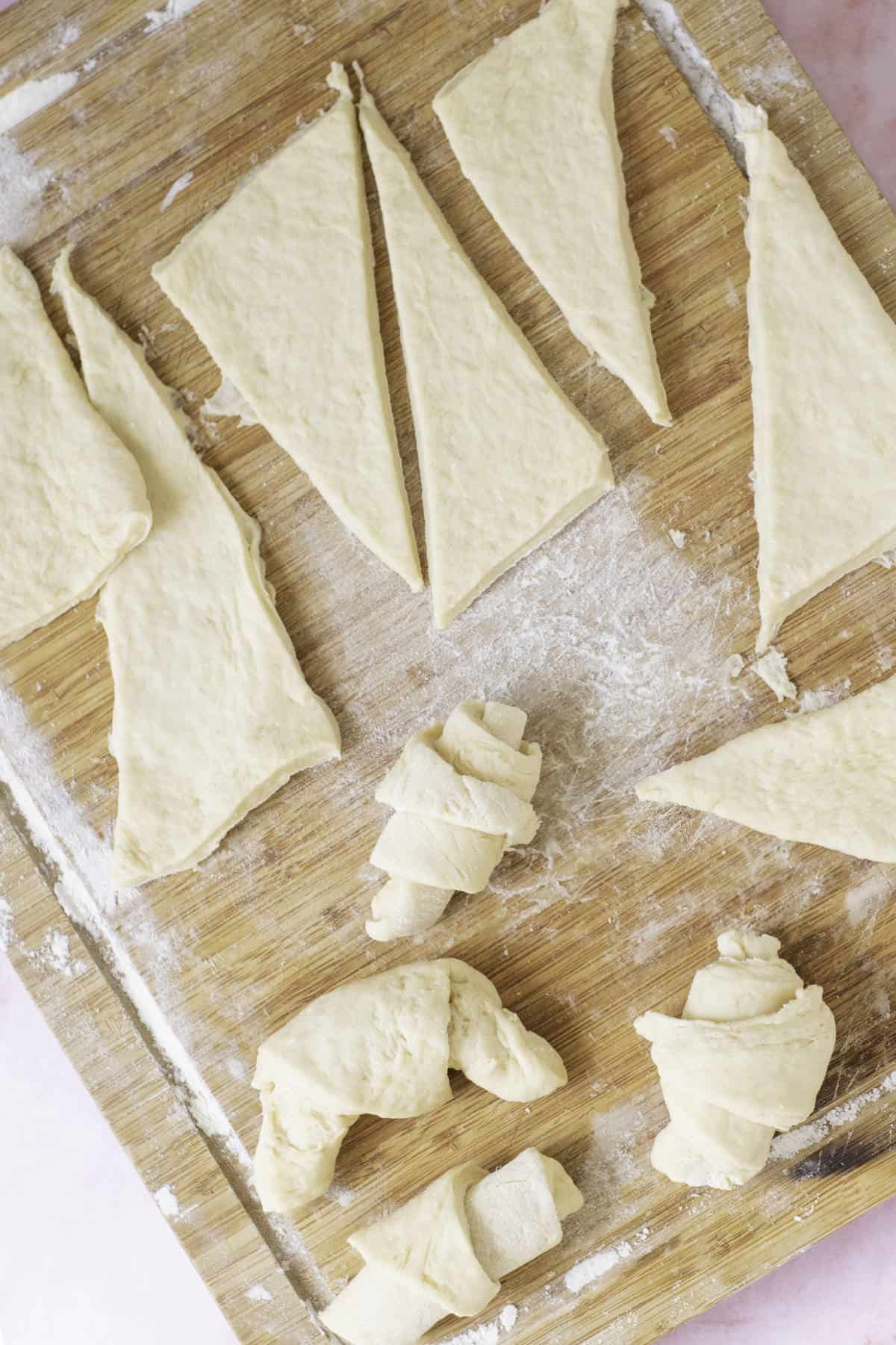 On a wooden board, unbaked homemade croissant dough rests, with some pieces expertly folded into croissant shapes and others in neat triangular cuts, flour generously scattered on the surface.