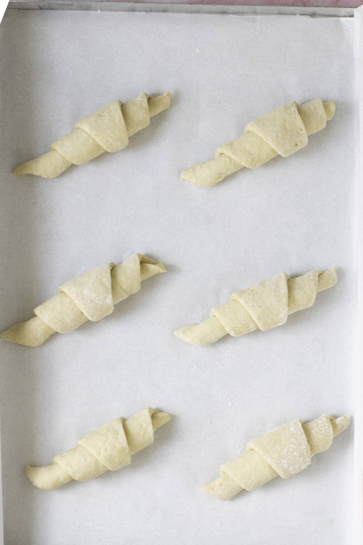Six fool-proof croissant dough pieces are evenly spaced on a parchment-lined baking tray. Each homemade croissant is rolled into a perfect crescent shape, ready for baking. The background is a light gray, adding contrast to the pale dough.