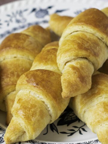 A plate with homemade croissants arranged closely. The fool-proof croissants have a golden-brown, flaky crust and are placed on a white plate with a blue floral pattern.