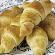 Close-up of freshly baked, golden-brown homemade croissants piled on a decorative white and blue plate. The croissants have a flaky texture and are arranged in an appetizing display.