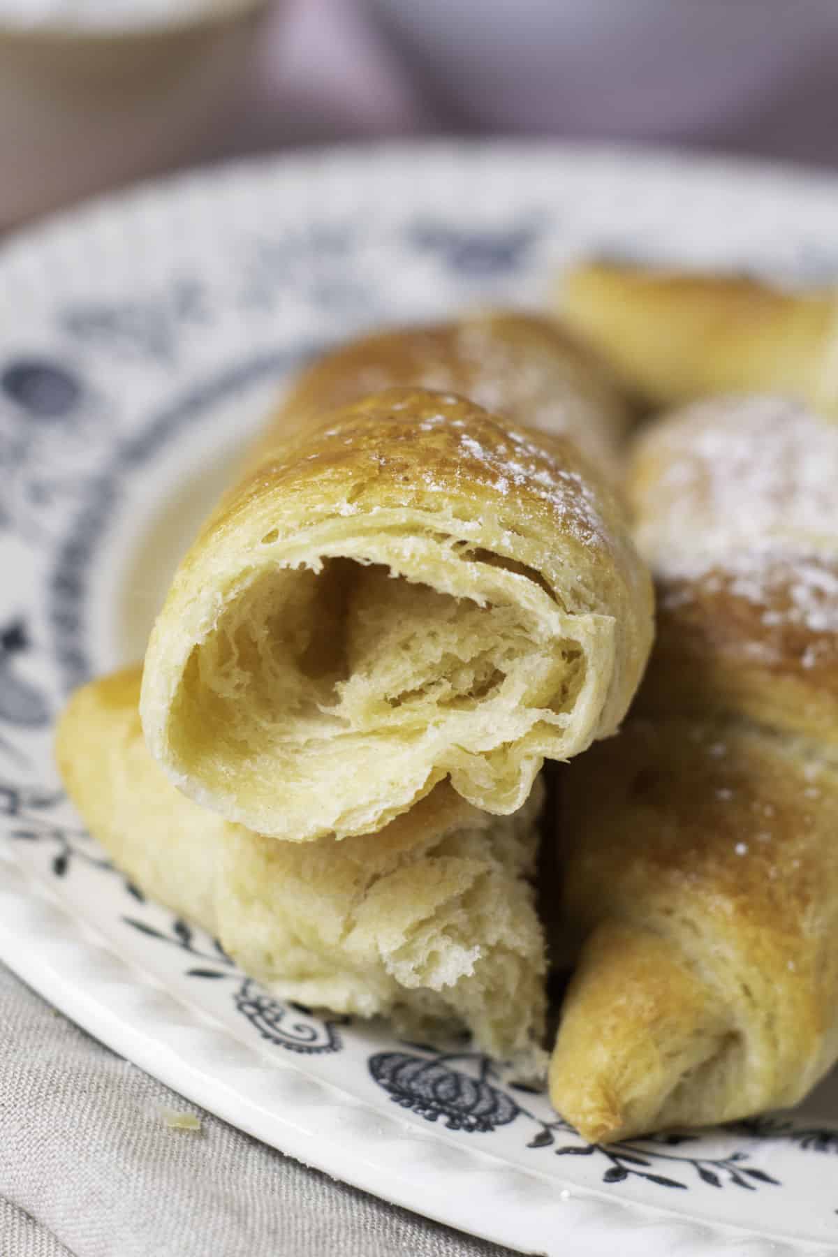 A close-up of buttery, golden-brown croissants on a decorative plate showcases their homemade charm. One fool-proof croissant is split open, revealing its flaky, layered interior. A light dusting of powdered sugar graces a nearby croissant.