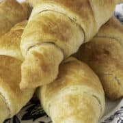 A plate with several fool-proof, freshly baked homemade croissants sits on a decorative floral-patterned dish. The golden-brown croissants are stacked and appear delightfully flaky and crisp.