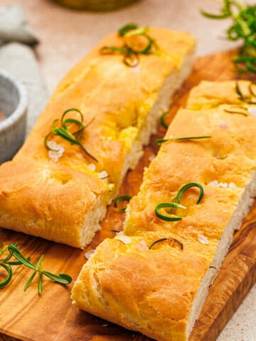 Freshly baked rosemary focaccia, sliced and garnished with fragrant sprigs and coarse salt, rests on a wooden cutting board. A small bowl of salt is nearby, and additional rosemary lays scattered for decoration.