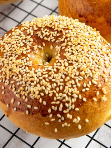 A freshly baked sesame bagel sits on a cooling rack, its golden brown crust adorned with sesame seeds and boasting a smooth, glossy surface.
