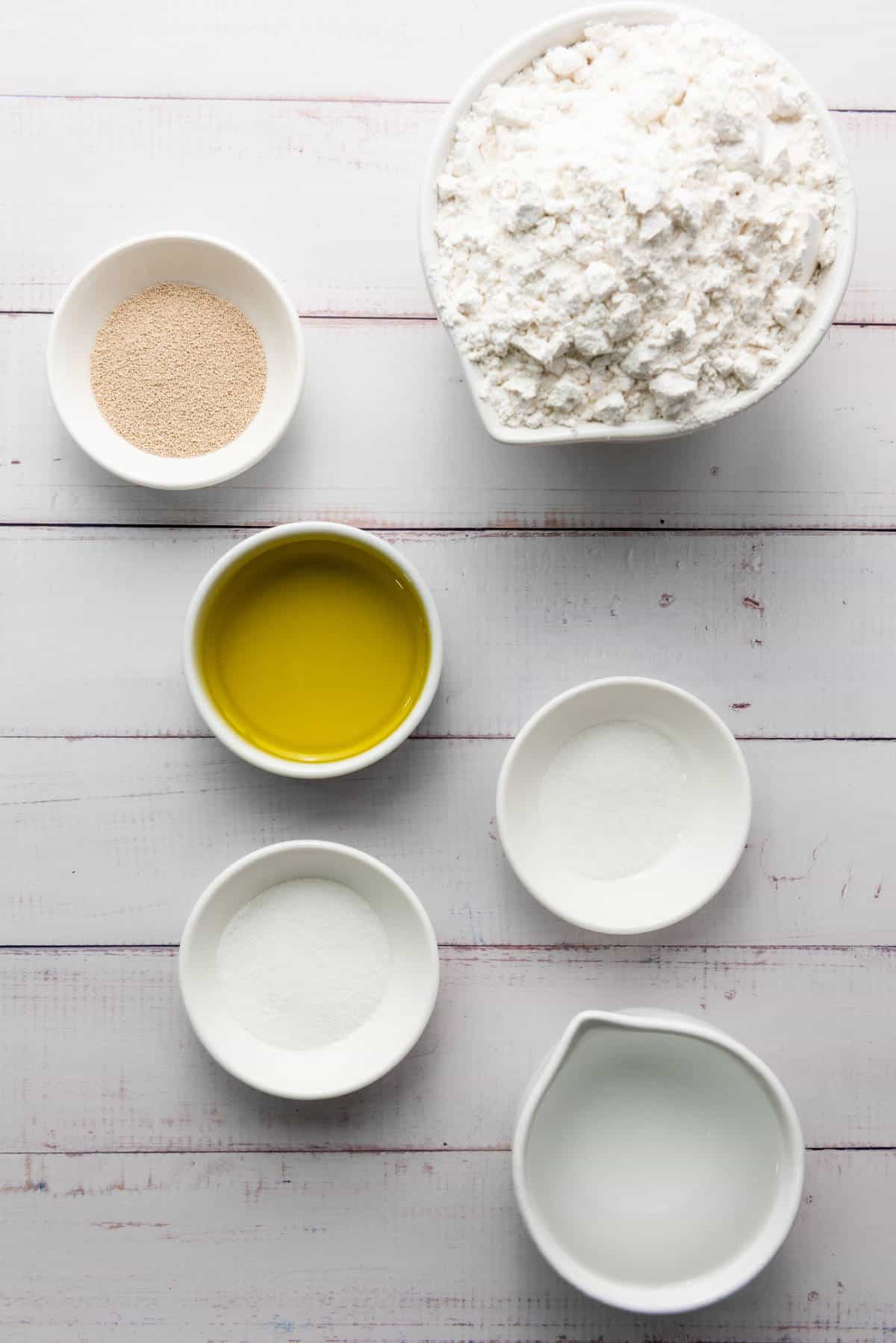 Ingredients for baking a thick crust pizza dough are arranged on a white wooden surface. This includes a bowl of flour, a small bowl of dry yeast, and another of olive oil, alongside three small bowls filled with sugar, salt, and water.