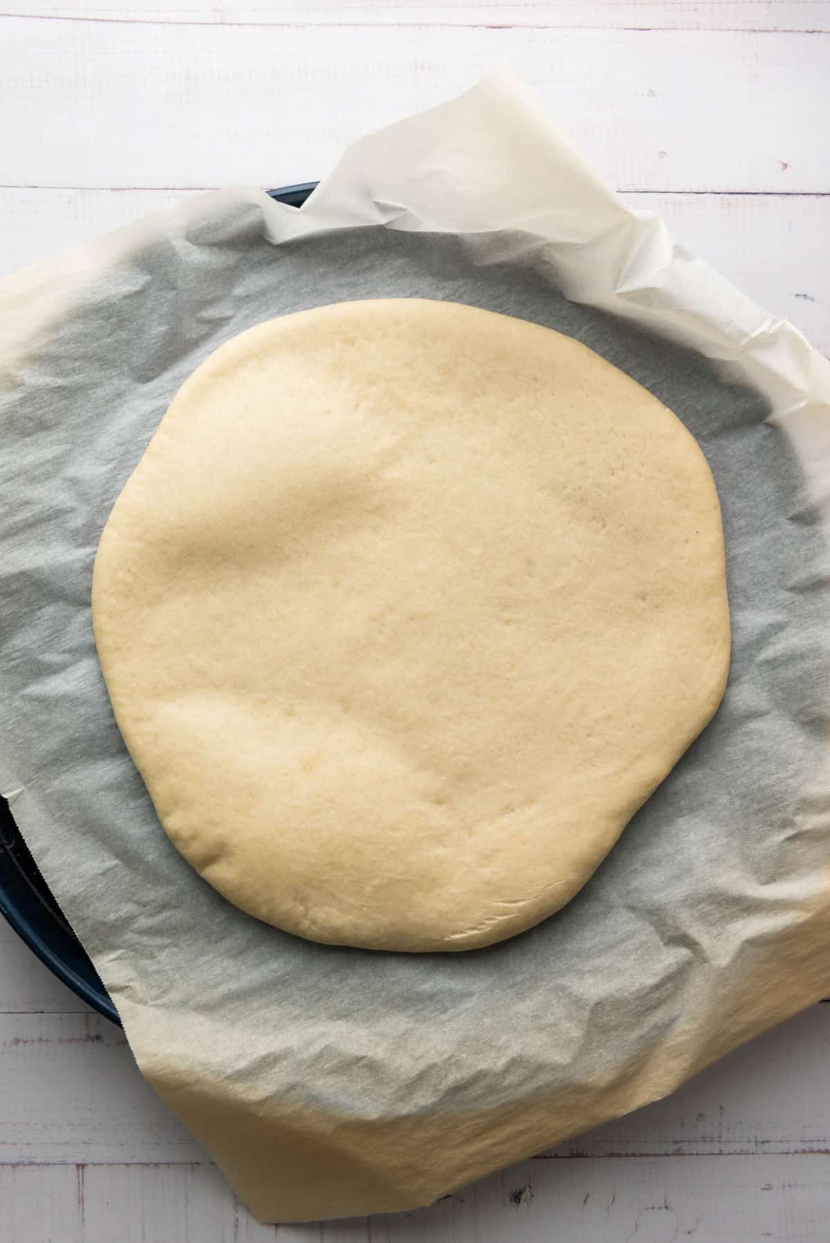 A thick crust pizza dough is placed on parchment paper, ready for baking, against a white wooden background.