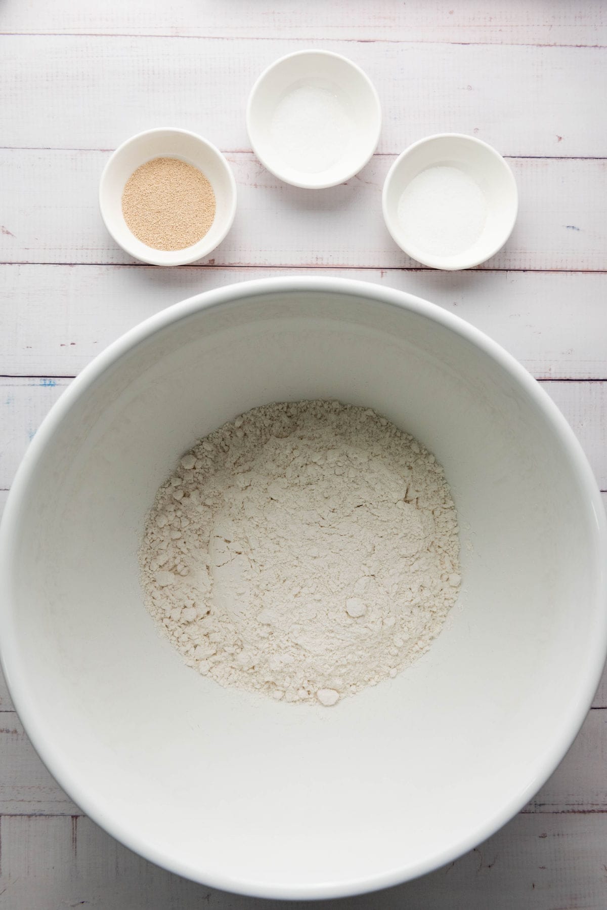 A white bowl containing flour is centered on a light wooden surface. Above it, three smaller bowls hold yeast, sugar, and salt-essential for crafting a thick-crust pizza dough. The ingredients are neatly arranged, suggesting preparation for baking.
