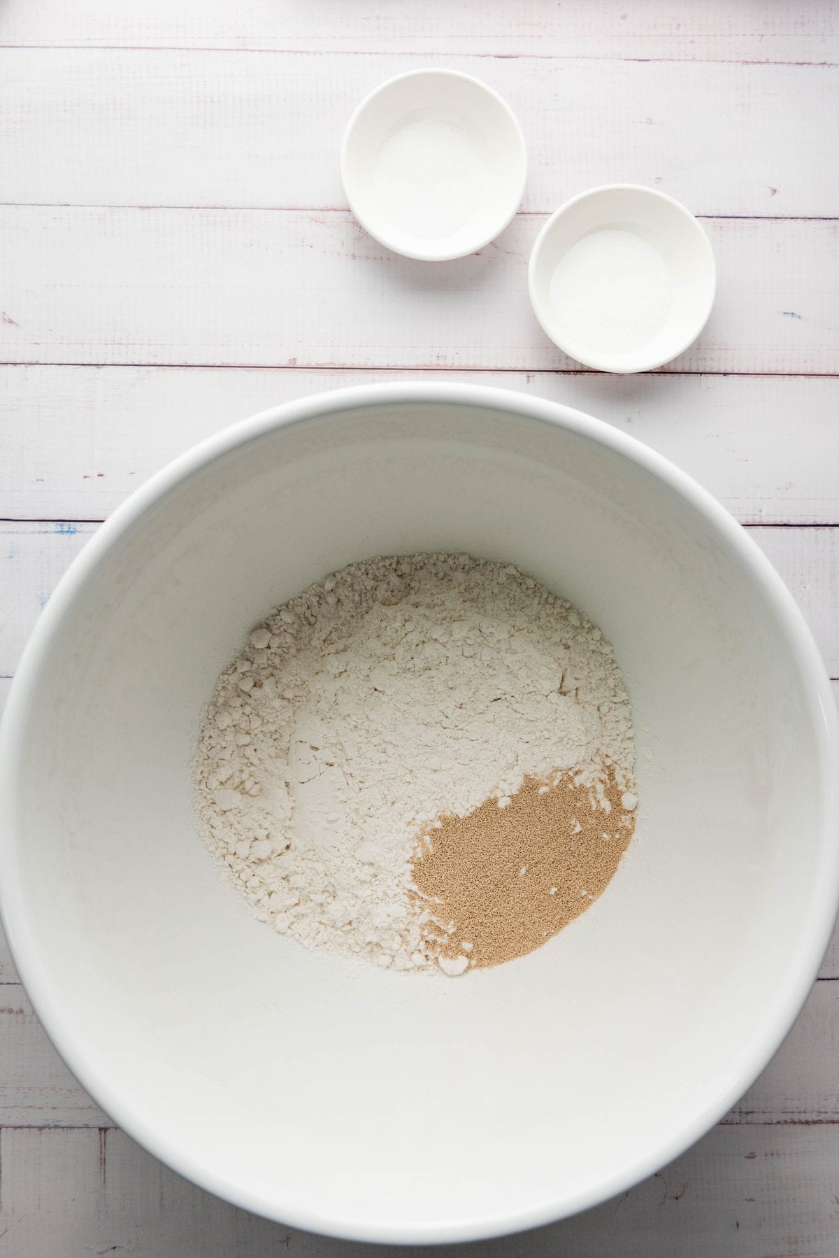 A large white bowl contains flour and yeast, perfect for making thick crust pizza dough, resting on a white wooden surface. Two small white bowls, one with salt and the other with sugar, are placed near the top of the image.