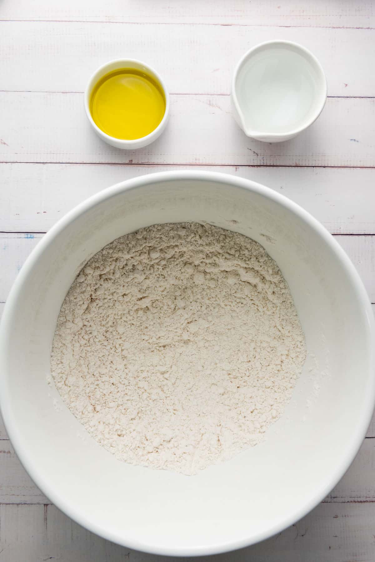 A large white bowl filled with flour sits on a white wooden surface, ready for crafting thick crust pizza dough. Next to it are two smaller bowls, one holding olive oil and the other water, essential ingredients for the perfect dough mix.