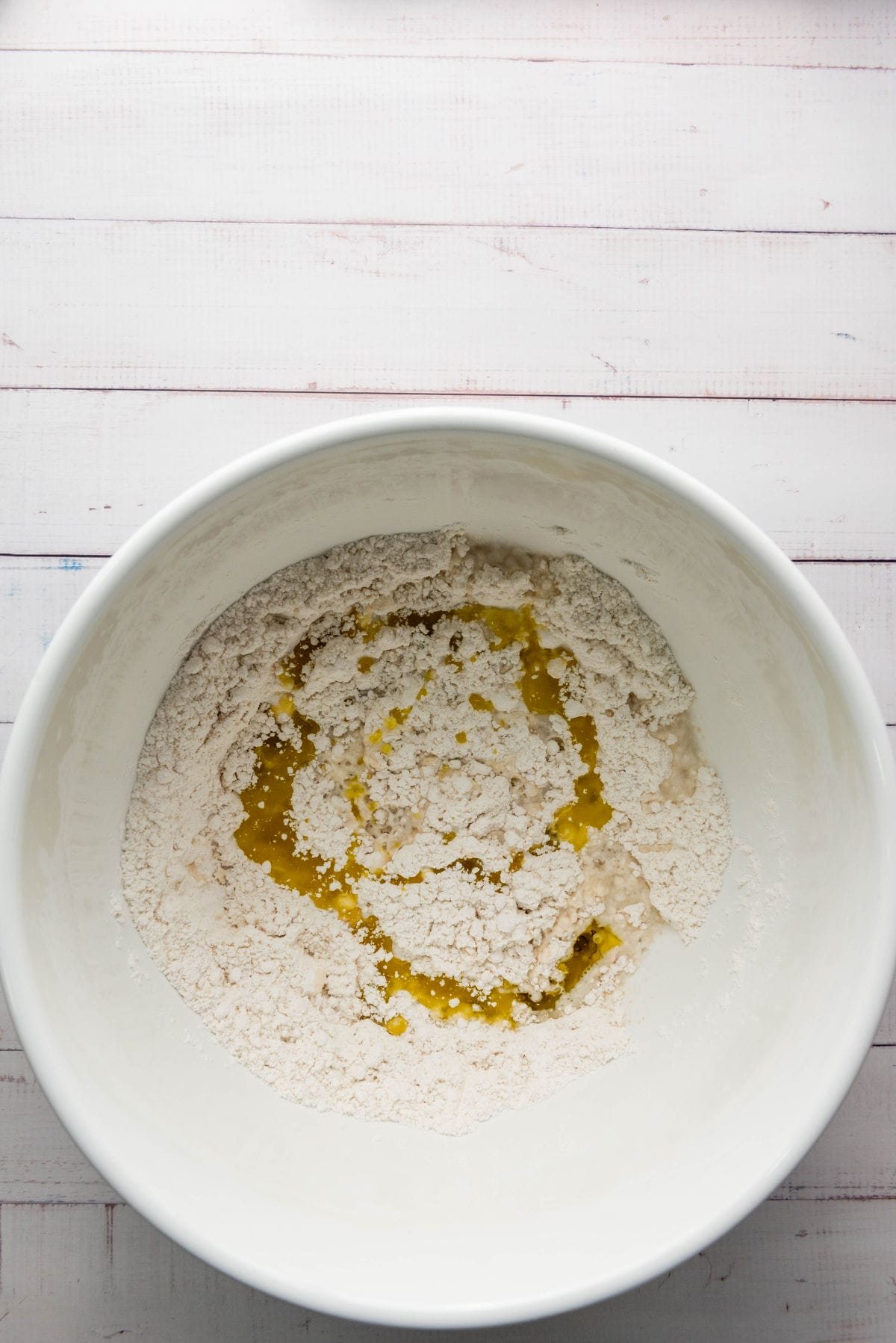 A white mixing bowl on a wooden surface contains flour and a pool of olive oil in the center, ready for mixing. The setting suggests preparation for crafting a thick crust pizza dough destined for an irresistible baking adventure.