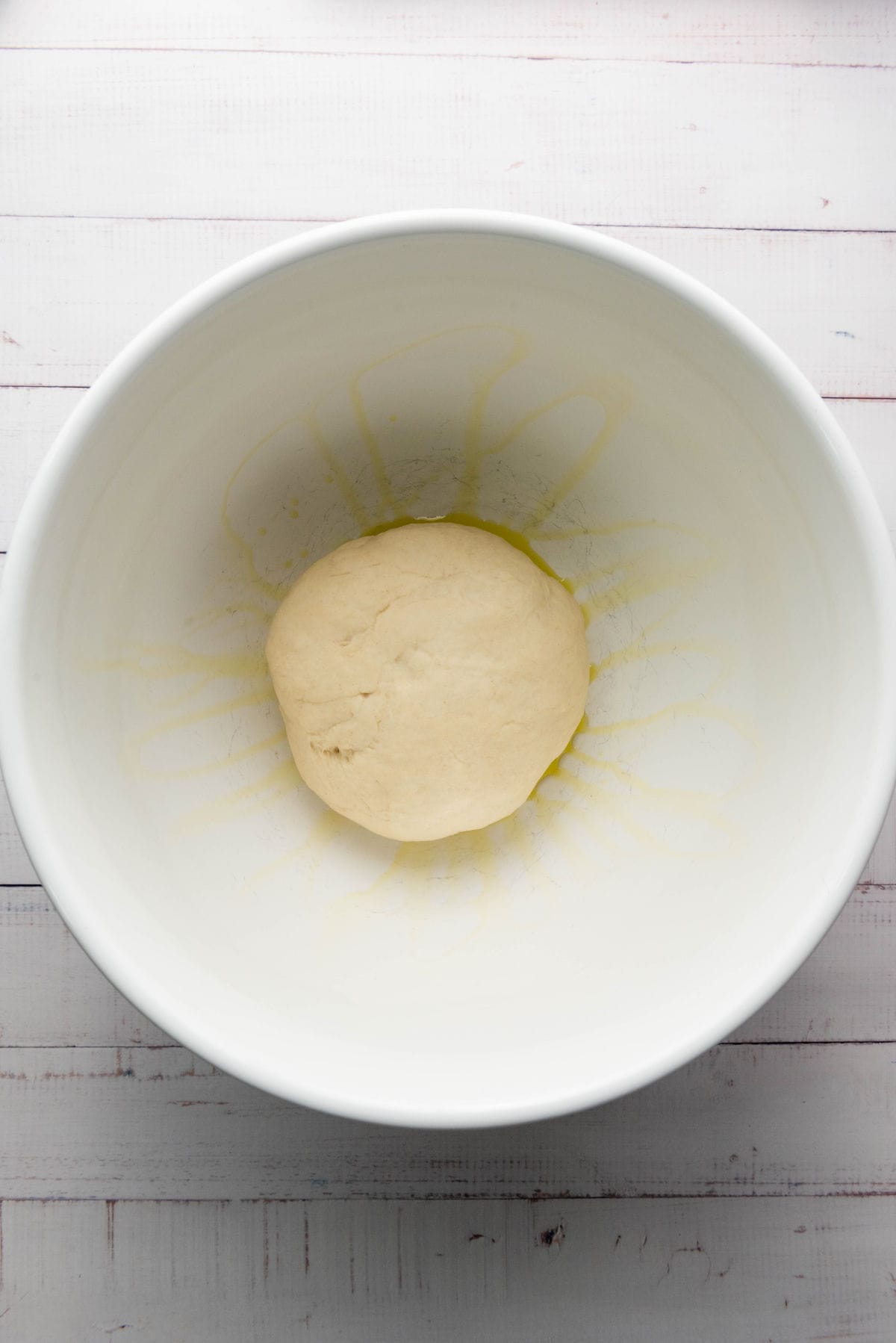 A ball of thick crust pizza dough rests in the center of a white mixing bowl, with a light coating of oil visible on the bottom and sides. The bowl is set on a white wooden surface.