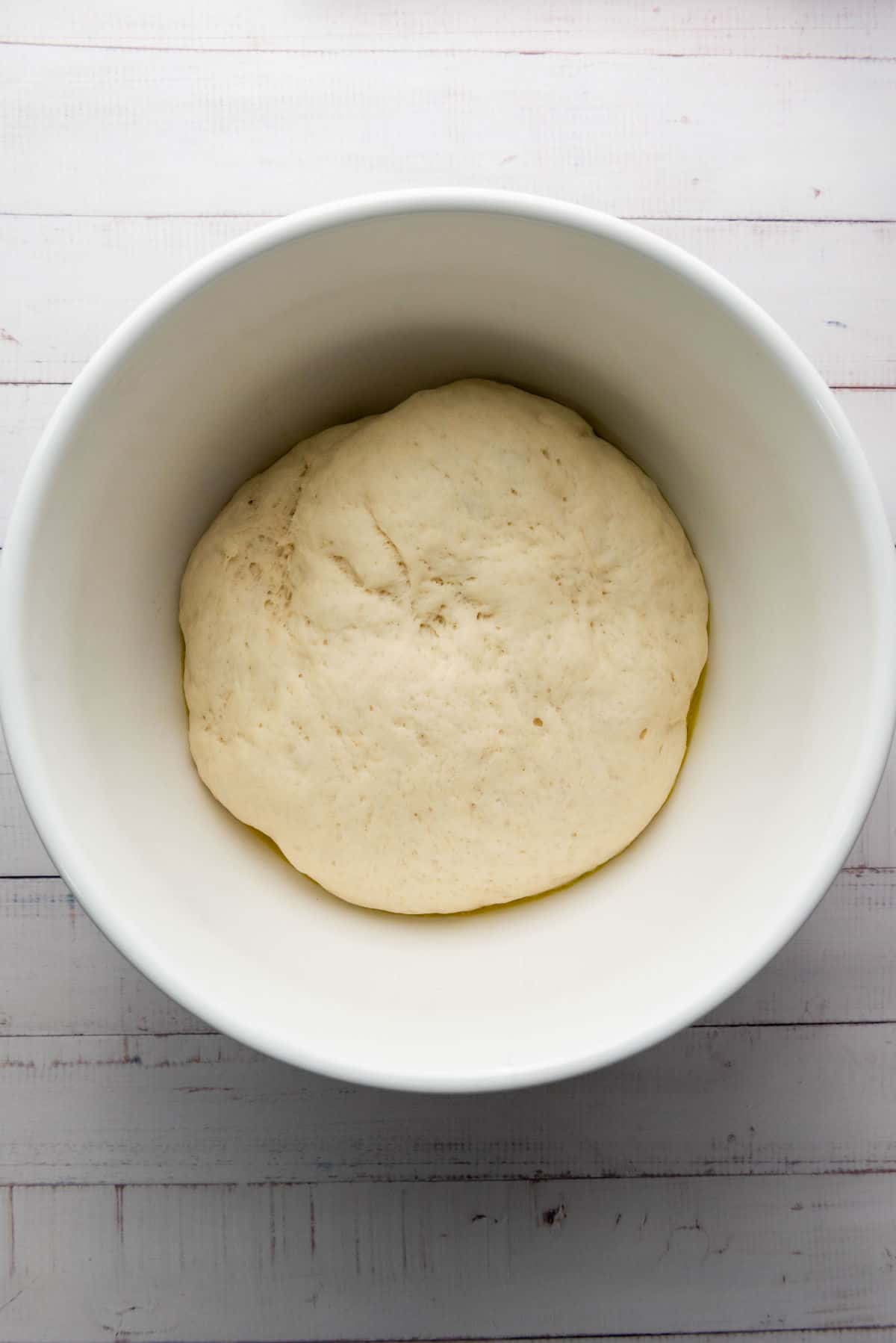 A ball of thick crust pizza dough resting in a white bowl on a light, textured surface. The dough appears smooth and slightly risen.
