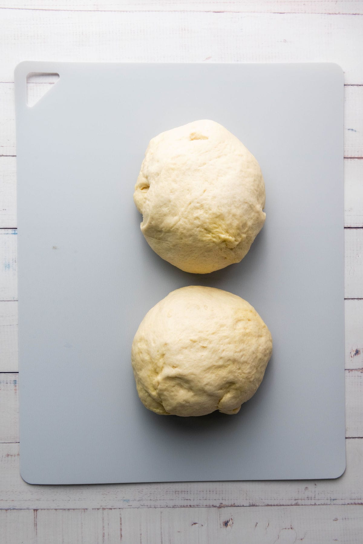 Two balls of thick crust pizza dough rest on a light-colored cutting board placed on a white wooden surface. The dough appears smooth and ready for baking preparations.