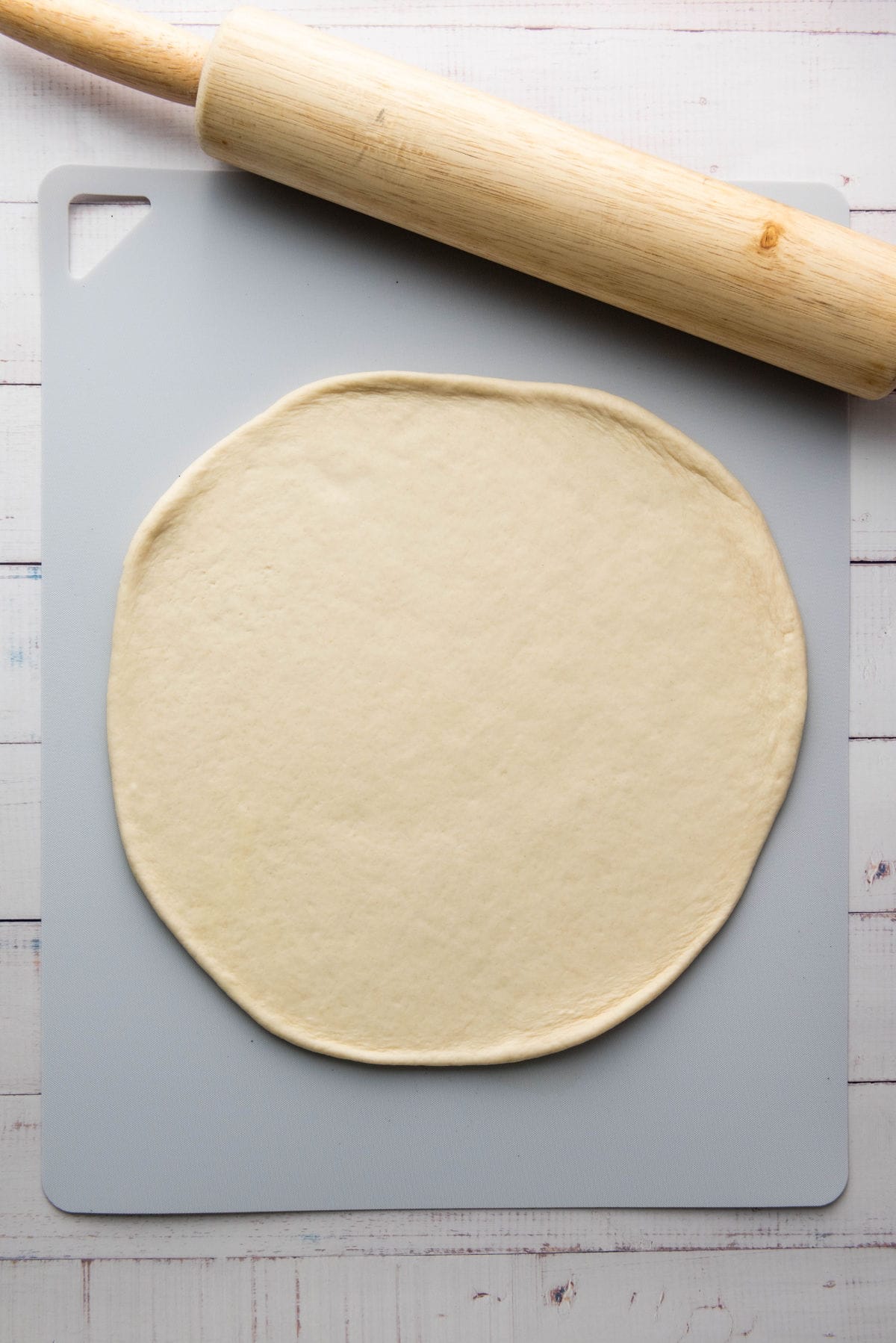 A flattened round of thick crust pizza dough rests on a gray cutting board with a wooden rolling pin placed diagonally atop. The setup is on a light-colored wooden table.