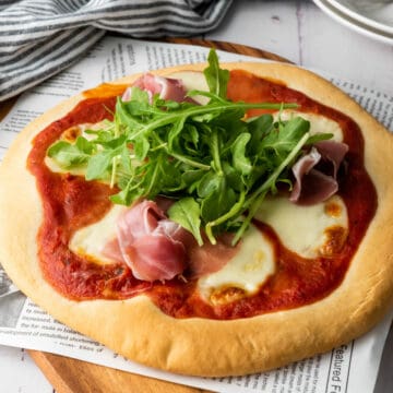 A thick crust pizza dough forms the base for this creation, topped with melted mozzarella, tomato sauce, prosciutto, and fresh arugula. Served on a wooden board partially covered with a newspaper, it sits alongside a striped cloth backdrop.