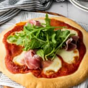 A freshly baked thick crust pizza topped with melted mozzarella, thin slices of prosciutto, and a heap of fresh arugula sits on a wooden board. A striped cloth and stacked plates with utensils are in the background.