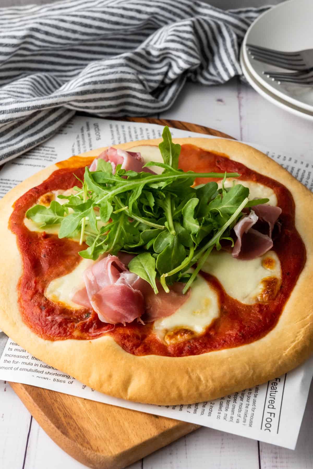 A freshly baked thick crust pizza topped with melted mozzarella, thin slices of prosciutto, and a heap of fresh arugula sits on a wooden board. A striped cloth and stacked plates with utensils are in the background.