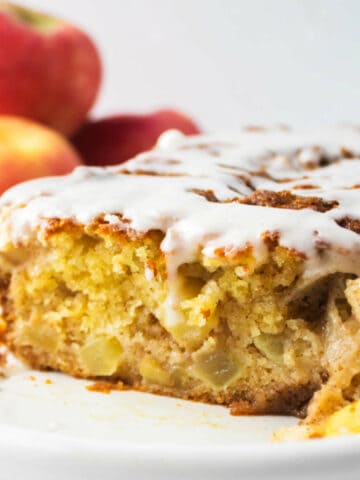 A close-up of a moist country apple fritter bread with chunks of apple inside, topped with a white glaze. There are whole apples blurred in the background.
