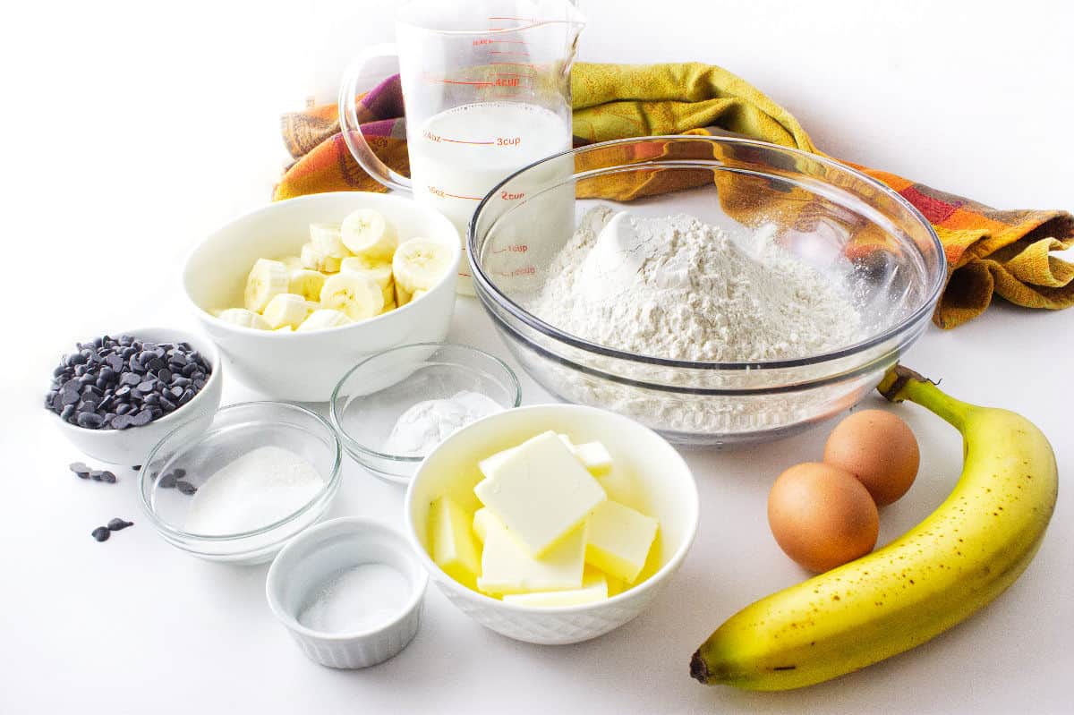 A collection of baking ingredients for banana chocolate chip sheet pancakes on a white surface, including flour, sliced bananas, whole bananas, chocolate chips, butter, sugar, baking powder, salt, eggs, milk, and a colorful cloth in the background.