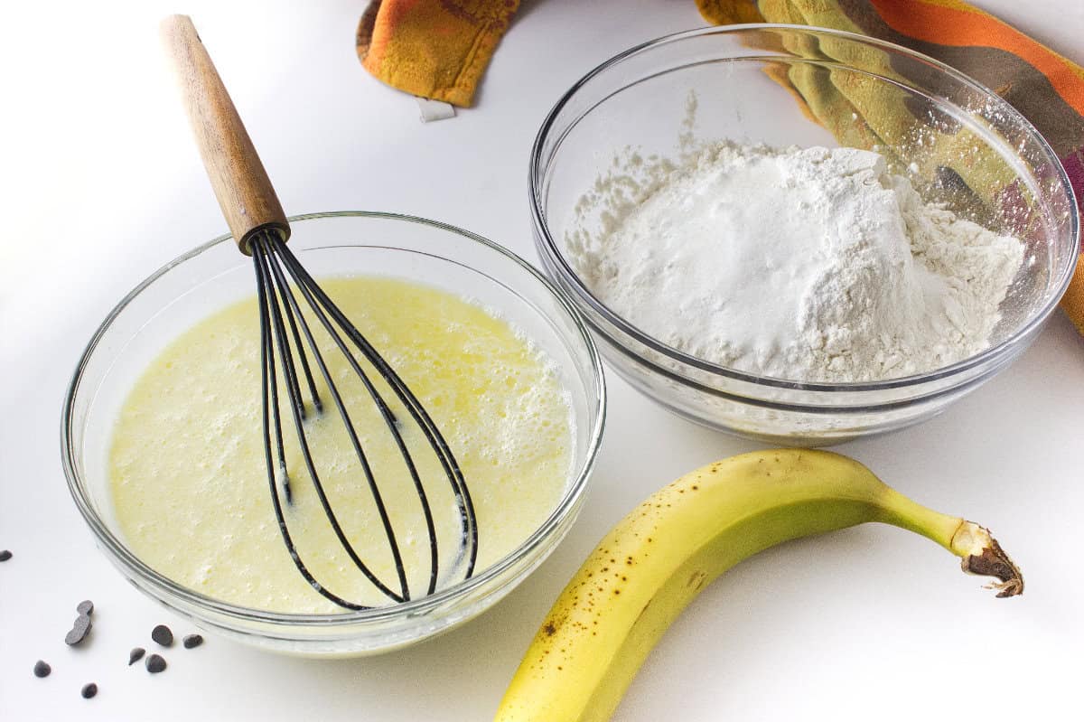Two glass bowls on a white surface: one with a whisk in a yellow liquid mixture for banana chocolate chip sheet pancakes, the other with flour. A ripe banana, chocolate chips, and a colorful cloth are nearby.