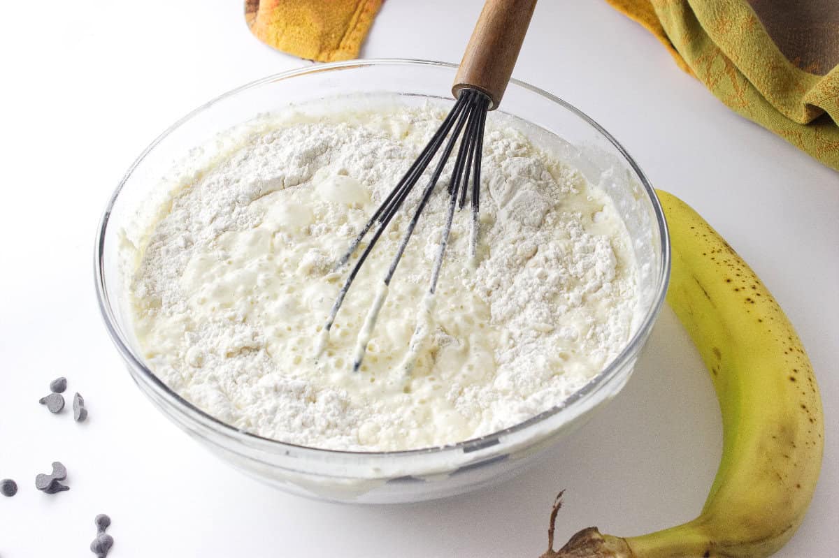 A glass bowl filled with flour, milk, and other banana chocolate chip sheet pancakes ingredients is being whisked. A ripe banana and a few chocolate chips rest on the white countertop, with a yellow kitchen towel in the background.