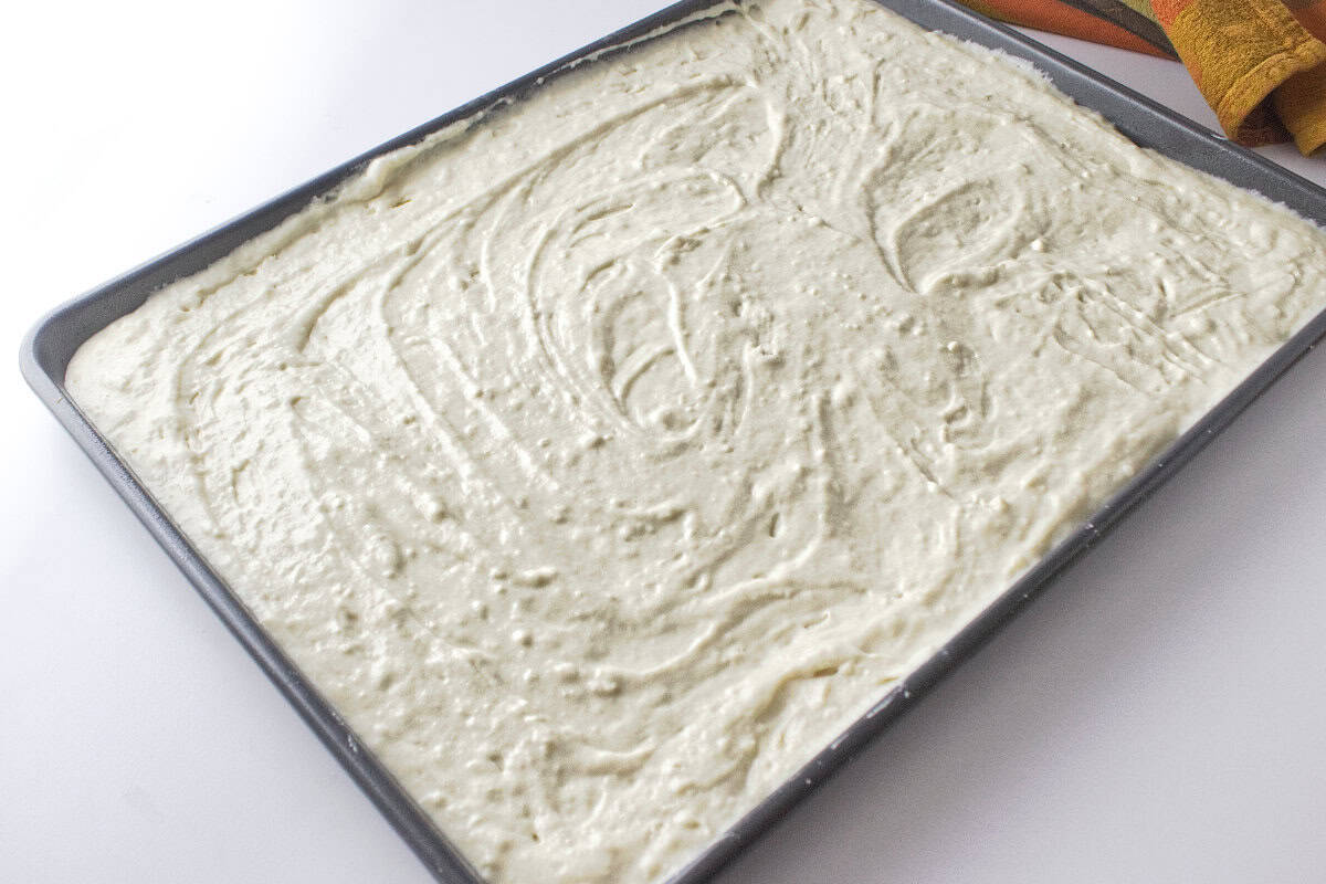 A baking sheet filled with raw banana chocolate chip sheet pancake batter evenly spread out, ready to be baked. The batter has a smooth, creamy texture with visible swirls and chocolate chips on the surface.