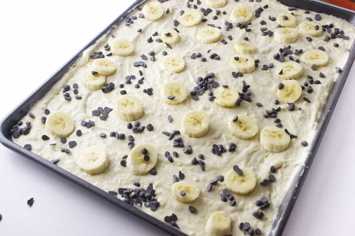 A baking sheet filled with banana chocolate chip sheet pancake batter, topped with banana slices and sprinkled with chocolate chips, ready to be baked.