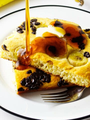 A plate with two square banana chocolate chip sheet pancakes topped with butter and syrup, a fork on the side, and a whole banana in the background.