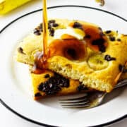 Two large pieces of banana chocolate chip sheet pancakes on a white plate, topped with butter and syrup being poured. A banana and chocolate chips are in the background; a fork rests beside the pancakes.