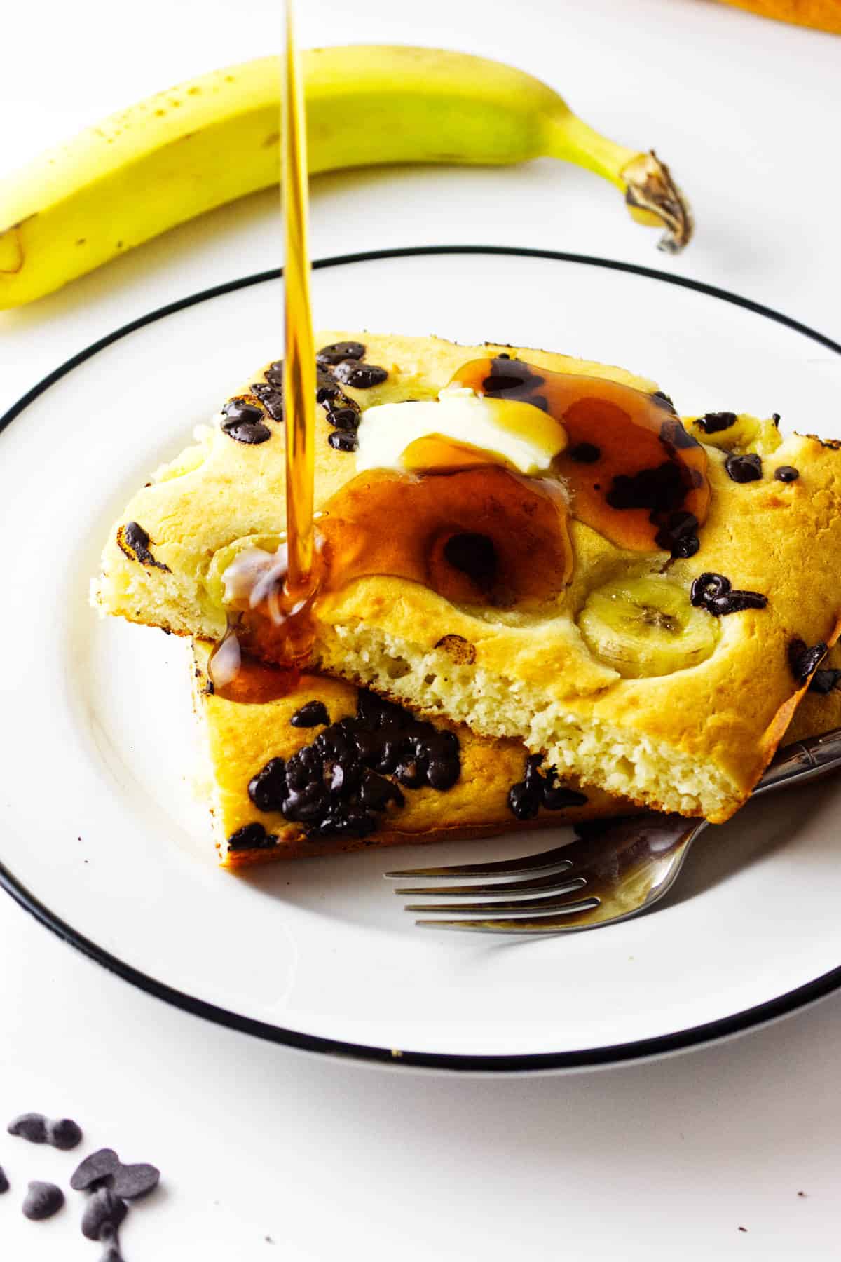 Two large pieces of banana chocolate chip sheet pancakes on a white plate, topped with butter and syrup being poured. A banana and chocolate chips are in the background; a fork rests beside the pancakes.