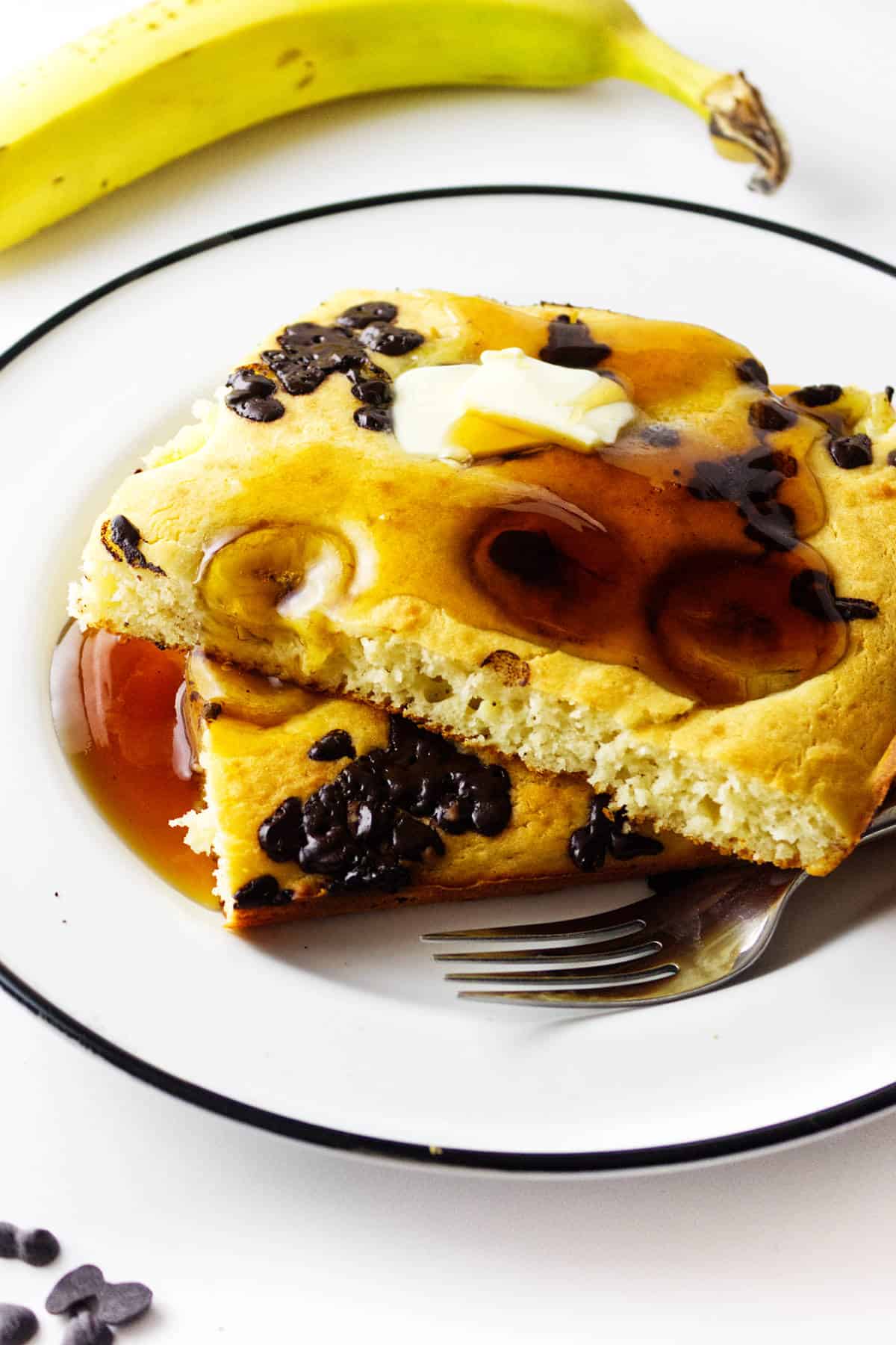A square of banana chocolate chip sheet pancakes topped with butter and syrup sits on a white plate with a fork. A banana is in the background, and a few chocolate chips are scattered nearby.