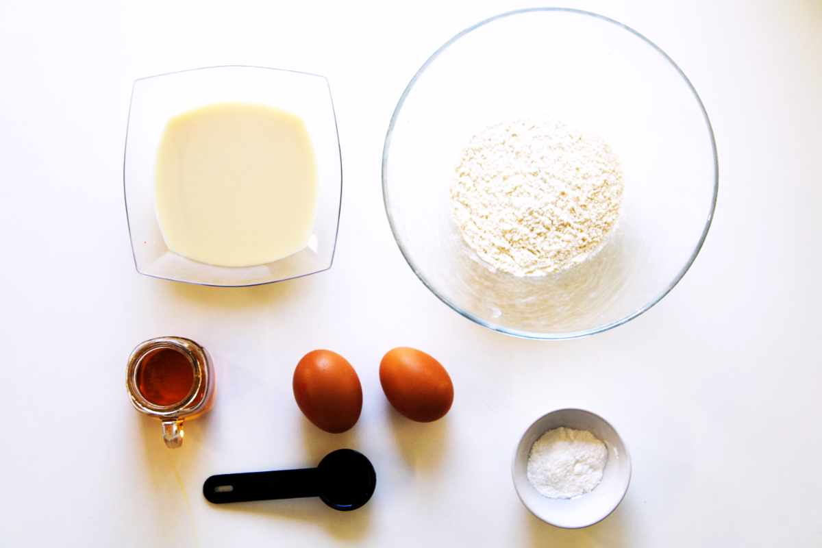 A top-down view of ingredients for baking old fashioned buttermilk pancakes: a bowl of flour, a bowl of milk, two brown eggs, a small cup of syrup, a black measuring spoon, and a small bowl of baking powder on a white surface.
