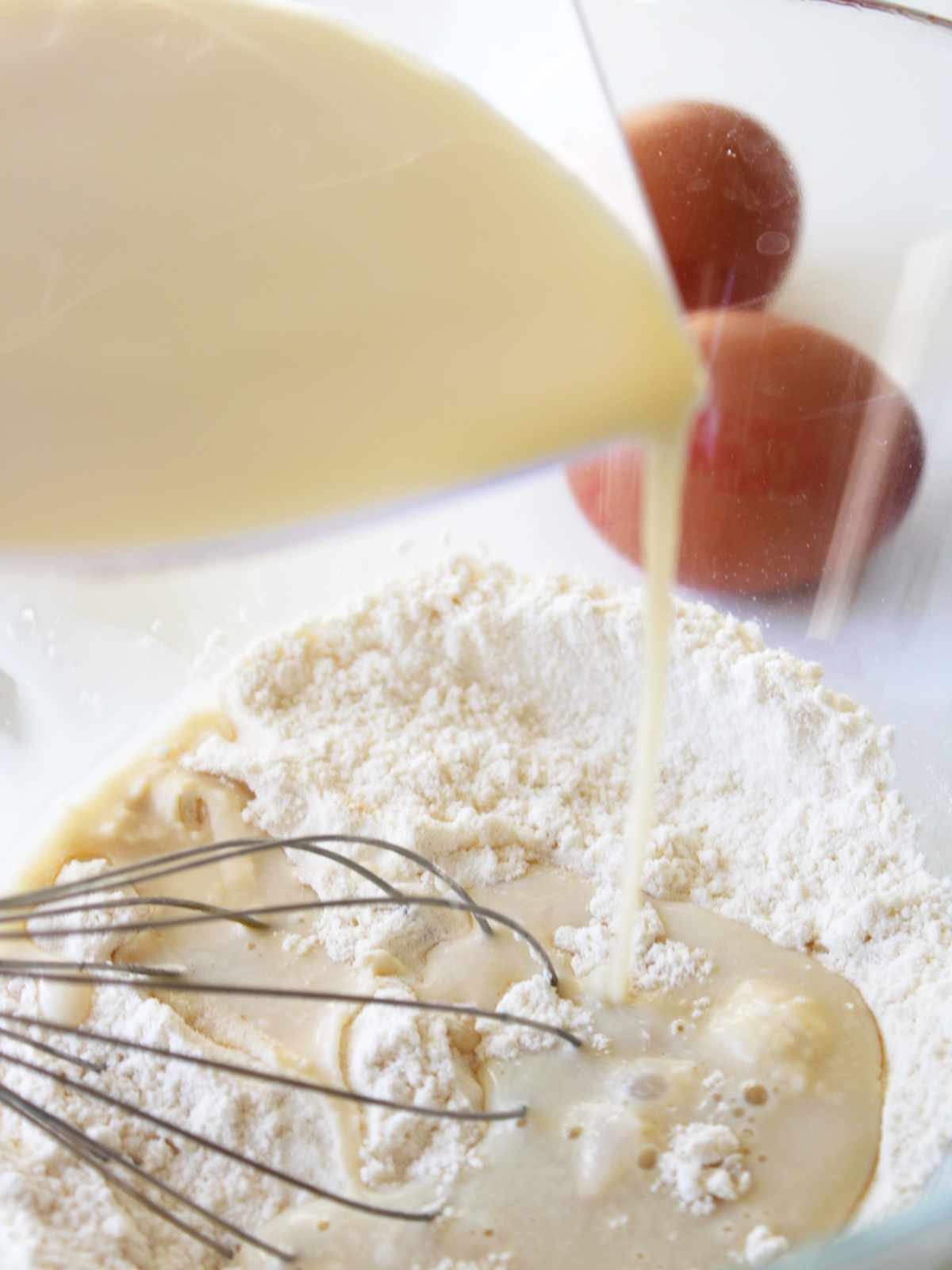 A close-up of milk being poured into a bowl of flour for old fashioned buttermilk pancakes, with a whisk in the mixture and two eggs in the background.