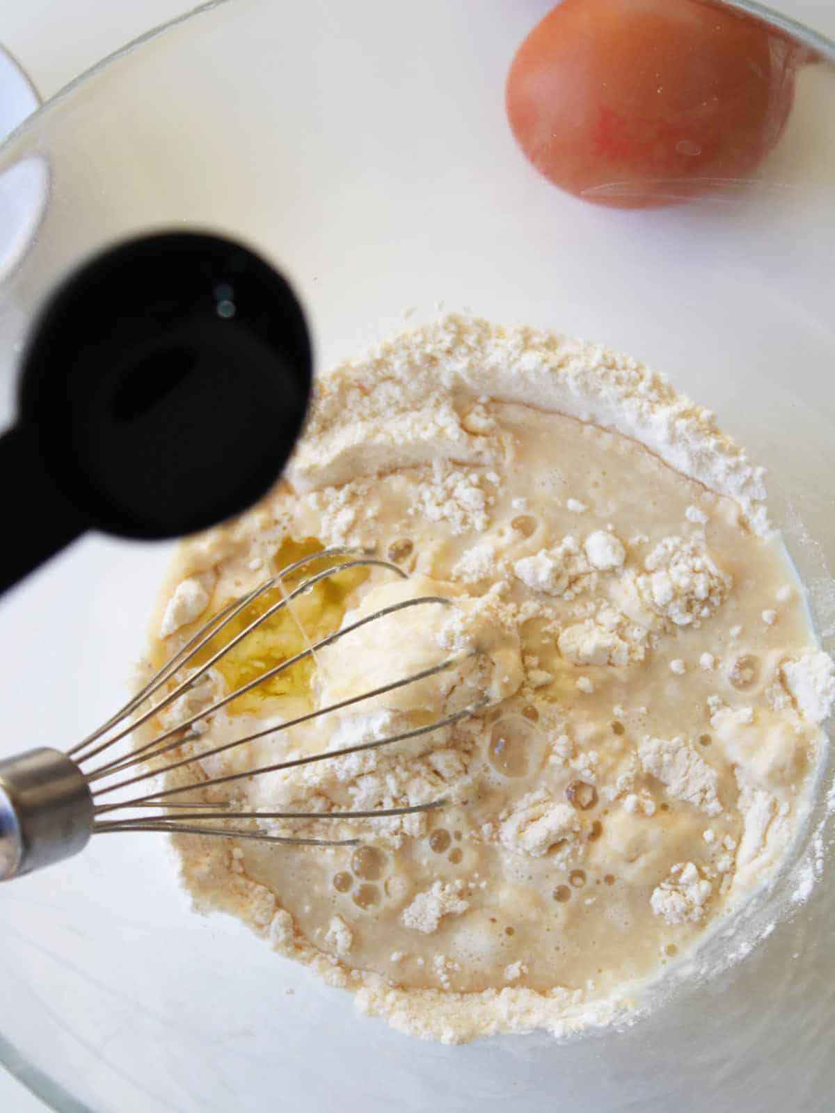 A glass bowl with flour, liquid ingredients, and a whisk inside—perfect for mixing old fashioned buttermilk pancakes. A measuring spoon hovers over the bowl, while an egg sits on the counter in the background.