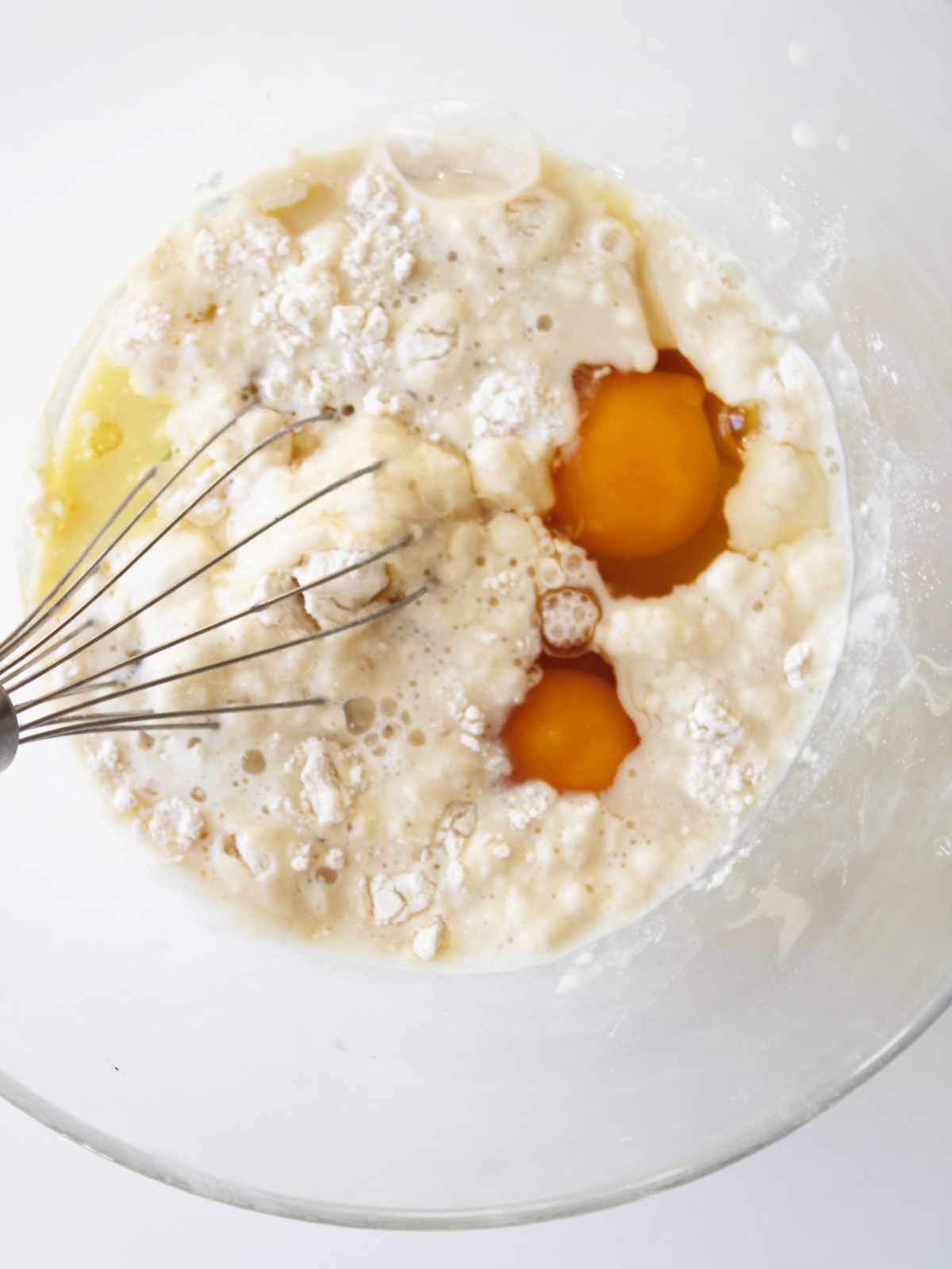 A whisk rests in a glass bowl containing eggs, flour, and milk, ready to be mixed together for baking a batch of old fashioned buttermilk pancakes.