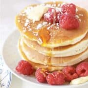 A stack of old fashioned buttermilk pancakes topped with fresh raspberries, a pat of butter, powdered sugar, and drizzled with syrup, served on a white plate with extra raspberries on the side.