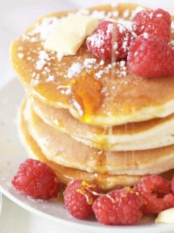 A stack of old fashioned buttermilk pancakes topped with fresh raspberries, a pat of butter, powdered sugar, and drizzled with syrup, served on a white plate with extra raspberries on the side.