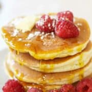 A stack of old fashioned buttermilk pancakes topped with fresh raspberries, a pat of butter, powdered sugar, and drizzled with syrup, with extra raspberries scattered on the plate.